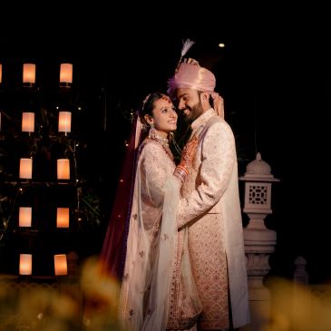 A newlywed couple embraces in traditional attire against a dark, candle-lit background.