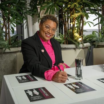 A woman in a black blazer and pink shirt signs documents at a table with plants in the background.