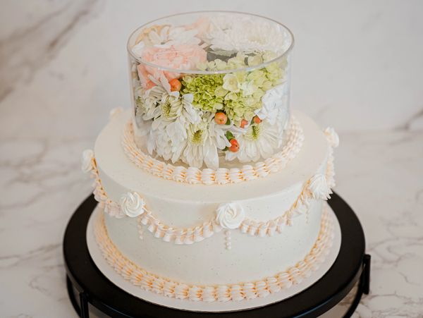 Elegant two-tier white cake with flowers encased in a transparent cylinder on top.