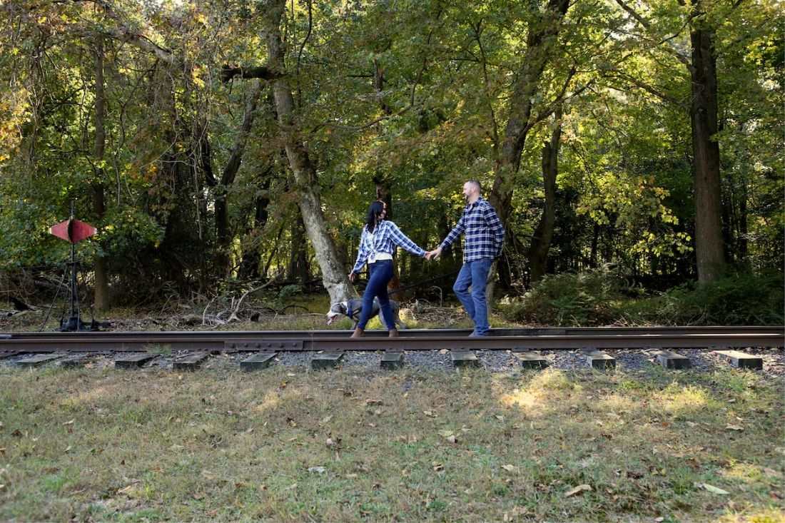 A cheerful dog being walked along train tracks by its two loving owners during their allaire state park engagement photo shoot.