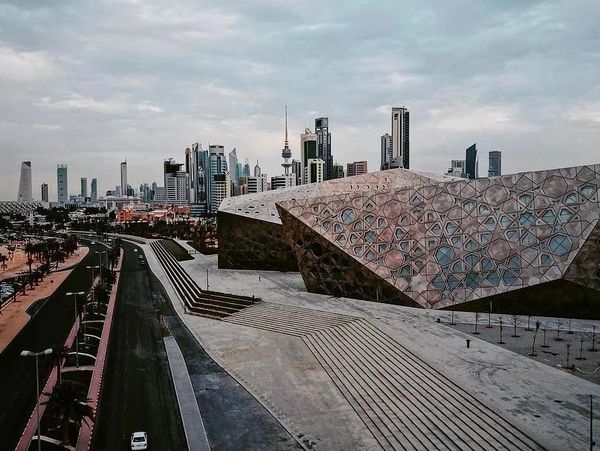 Modern geometric building with city skyline and road under cloudy sky.
