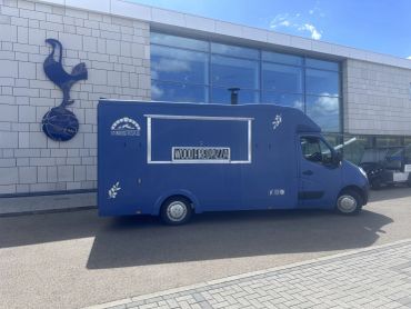 A blue food truck parked outside a modern building with a Tottenham Hotspur logo.