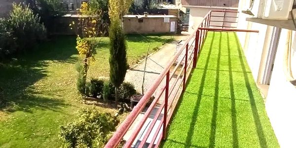 Balcony with green artificial grass and red railing overlooking a garden and houses.
