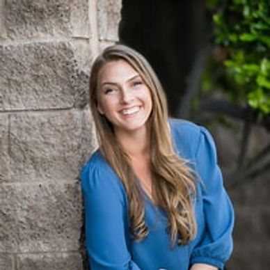 Smiling woman in a blue blouse leaning against a stone wall outdoors.