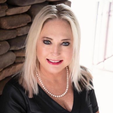 Smiling woman with blonde hair and pearl necklace indoors.