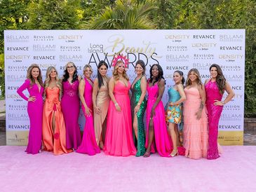 Group of women in elegant dresses posing on a pink carpet at a beauty awards event.