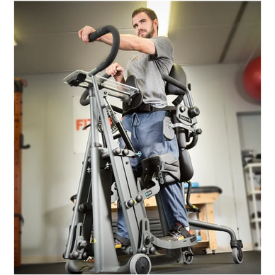 A Man in blue jeans and a grey t-shirt standing in an EasyStand Glier standing frame in a gym. 