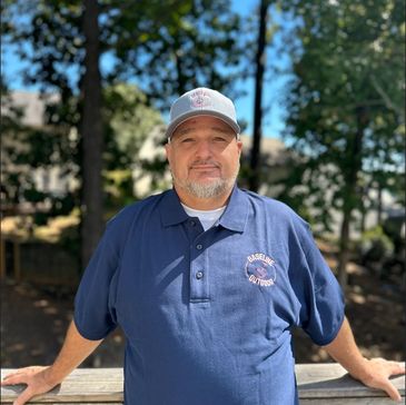 Man in a blue polo shirt and cap standing outdoors with trees in the background.