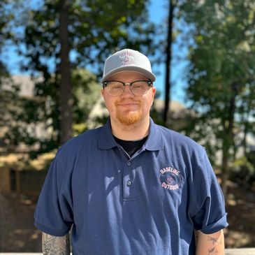 Man wearing glasses and a Baseline Outdoor cap and shirt, standing outdoors.