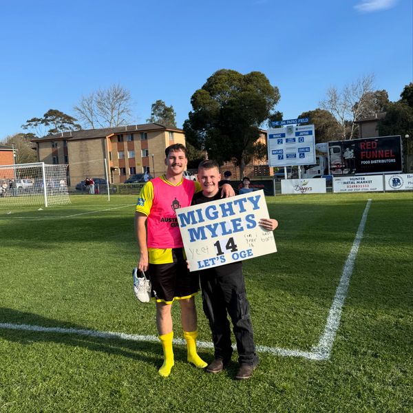 Two smiling boys on a soccer field holding a sign that says Mighty Myles 14 Let's OGE.
