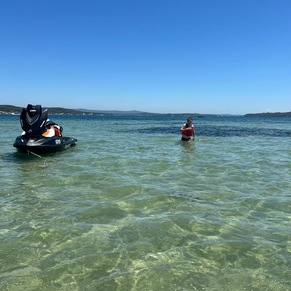 Person standing in clear shallow water near a jet ski on a sunny day.