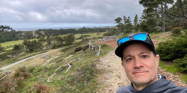 Man in a hoodie and cap taking a selfie on a scenic, cloudy hillside trail.