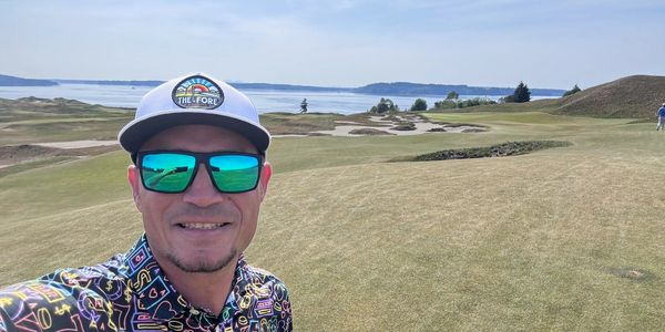 Man wearing reflective sunglasses and a colorful shirt on a golf course with water in the background.