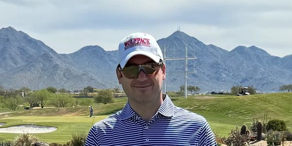 Man in striped shirt and cap on a golf course with mountains behind.