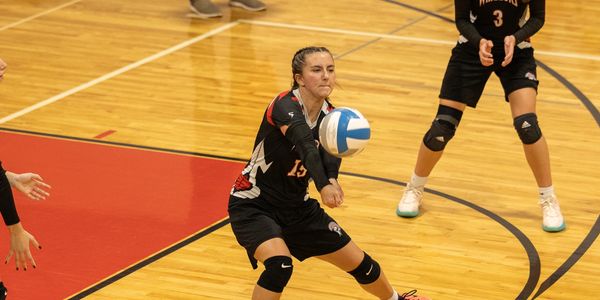 Female volleyball player in black uniform performing a dig on a gym court.