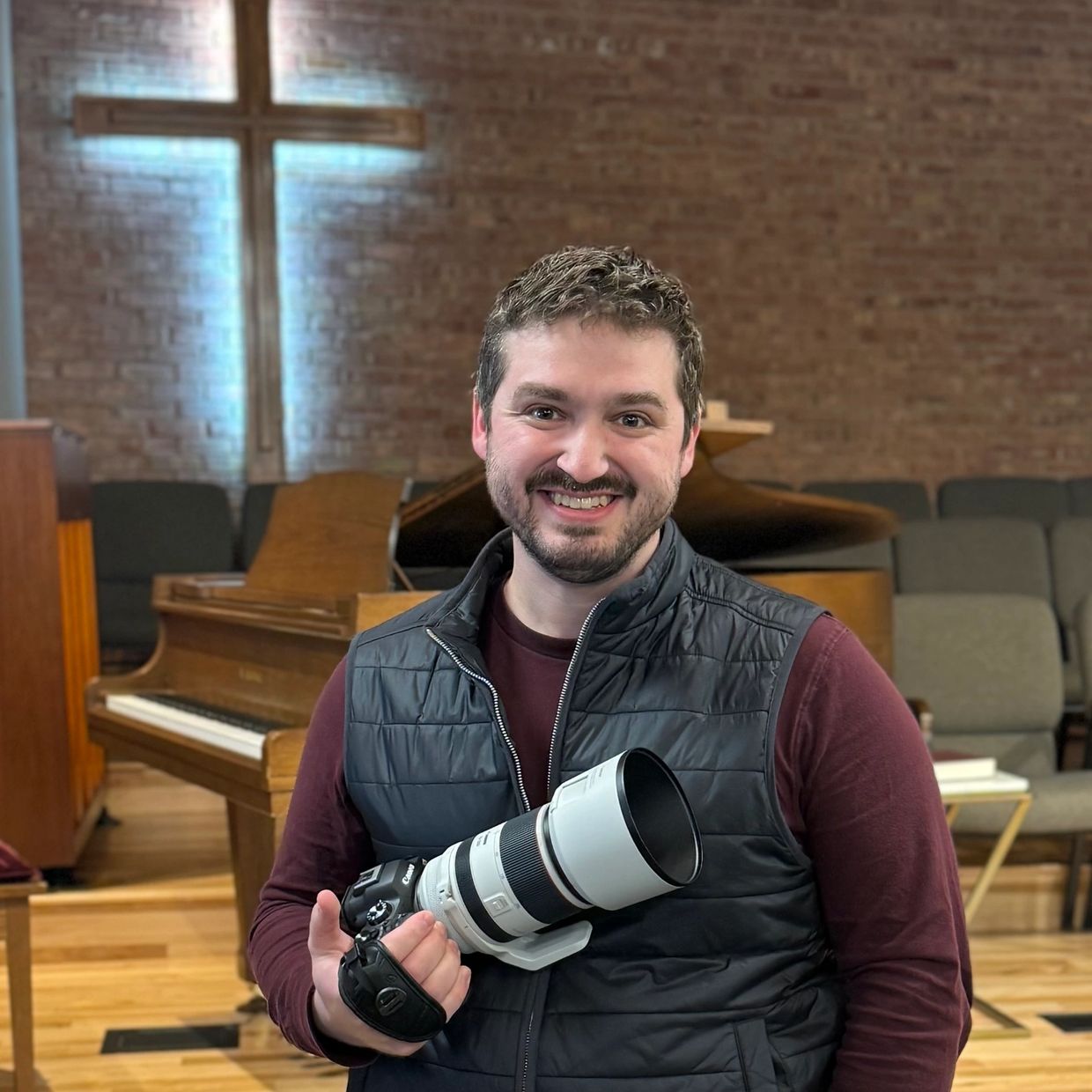 Smiling man holding a professional camera in a church setting with a piano and illuminated cross.