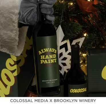 A gloved hand holding a Brooklyn Winery bottle labeled 'Always Hand Paint' near a festive Christmas tree.