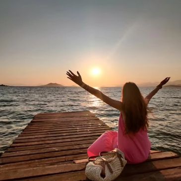 Woman sitting on a dock with arms raised, watching the sunset over the ocean.