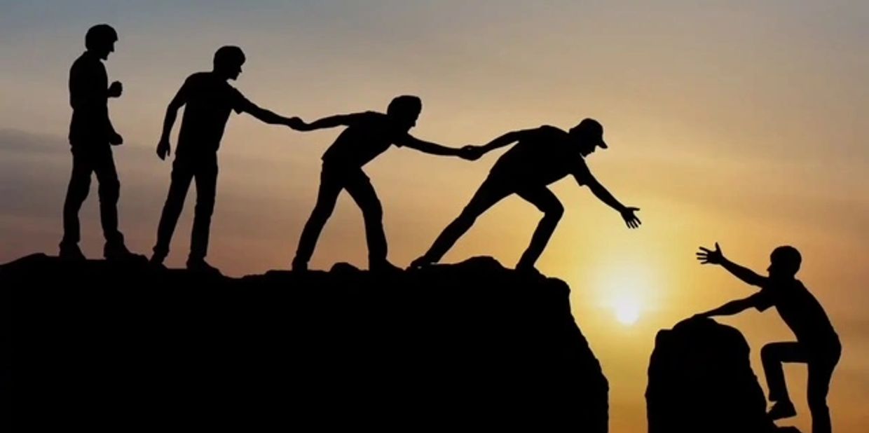 Silhouettes of friends helping each other climb rocks at sunset.
