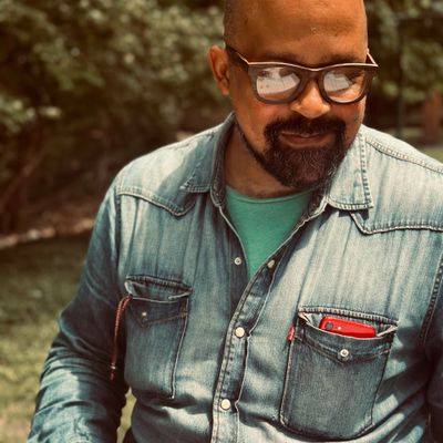 A man wearing glasses and a denim shirt outdoors, smiling.