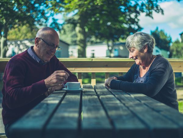 An older man and woman sit opposite each other at a picnic table with a hot drink