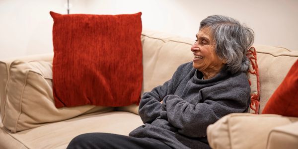 An older woman sits on a sofa surrounded with cushions, arms folded and smiling