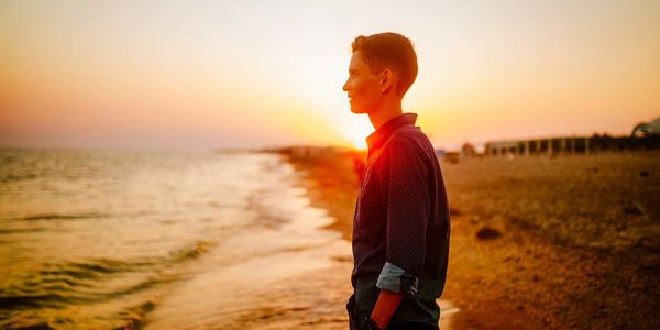 A young person stands on a beach looking out to sea with the sun setting behind them