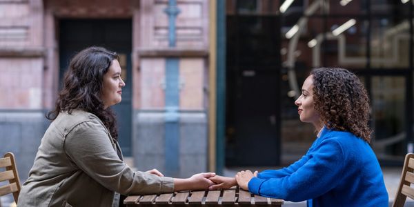 Two young women sit opposite each other, holding hands across an outdoor restaurant table