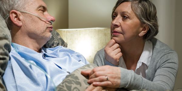 A man in bed with an oxygen tube in his nose holds hands with his wife who is next to the bedside