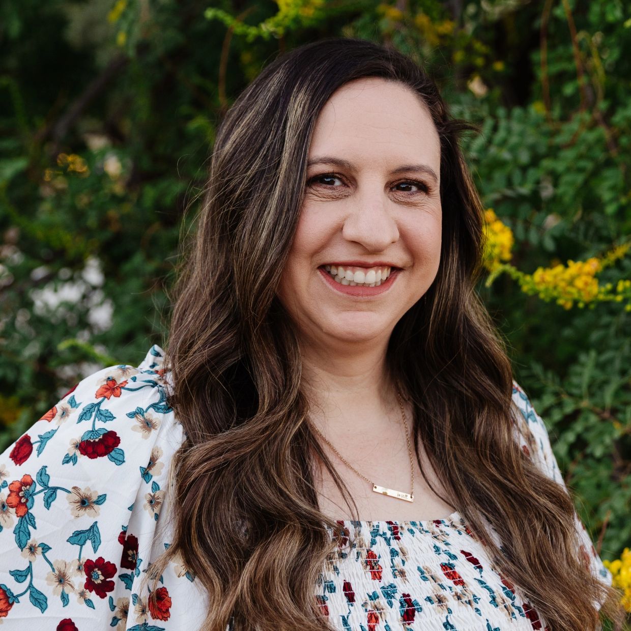 Smiling woman with long hair and floral top outdoors.