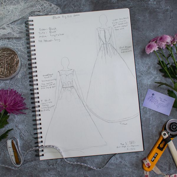 Pencil sketch of a wedding dress on a sketchbook surrounded by sewing equipment and pink flowers