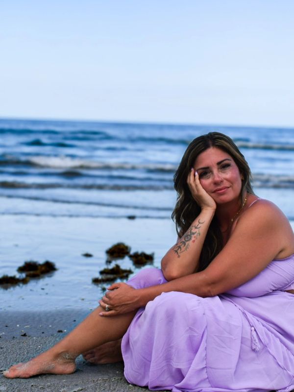 Woman in a lavender dress sitting on the beach, relaxing by the ocean.