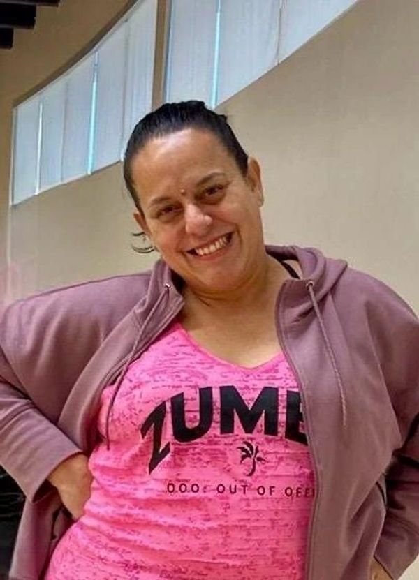 A smiling woman in a pink Zumba shirt and jacket indoors.