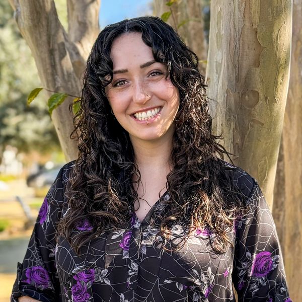 Woman smiling outdoors wearing a black shirt with purple flowers.