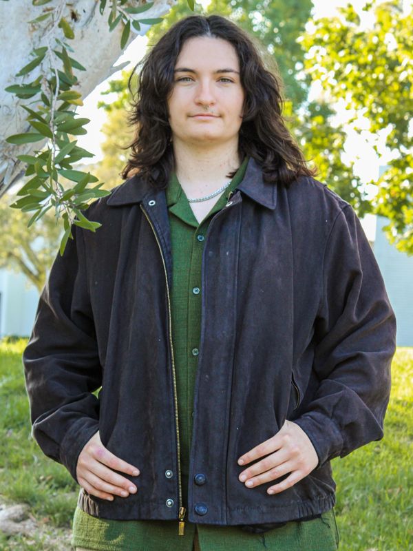 Person standing outdoors near a tree, wearing a dark jacket and green shirt.