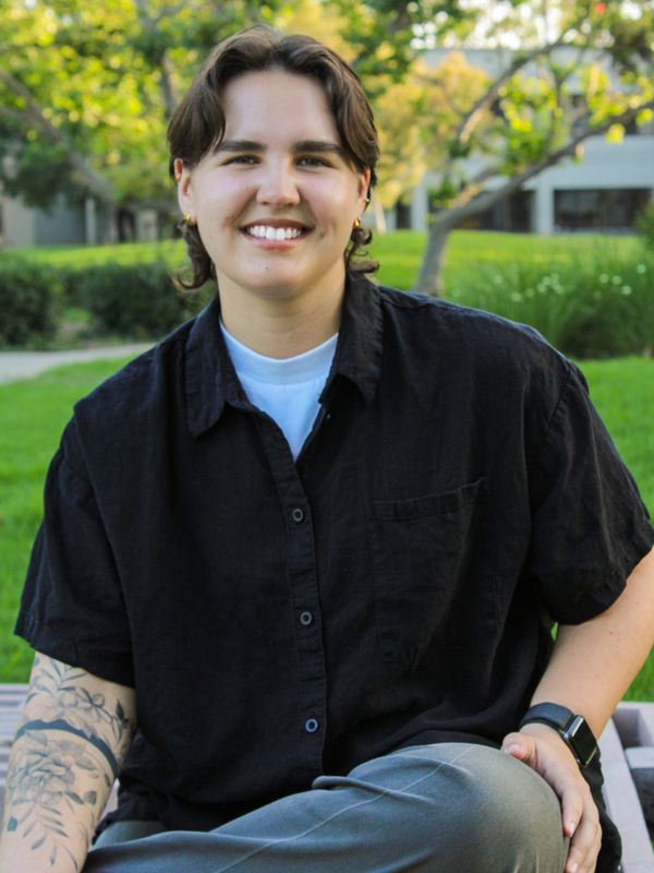 A smiling person with tattoos and short hair sitting outdoors on a bench.
