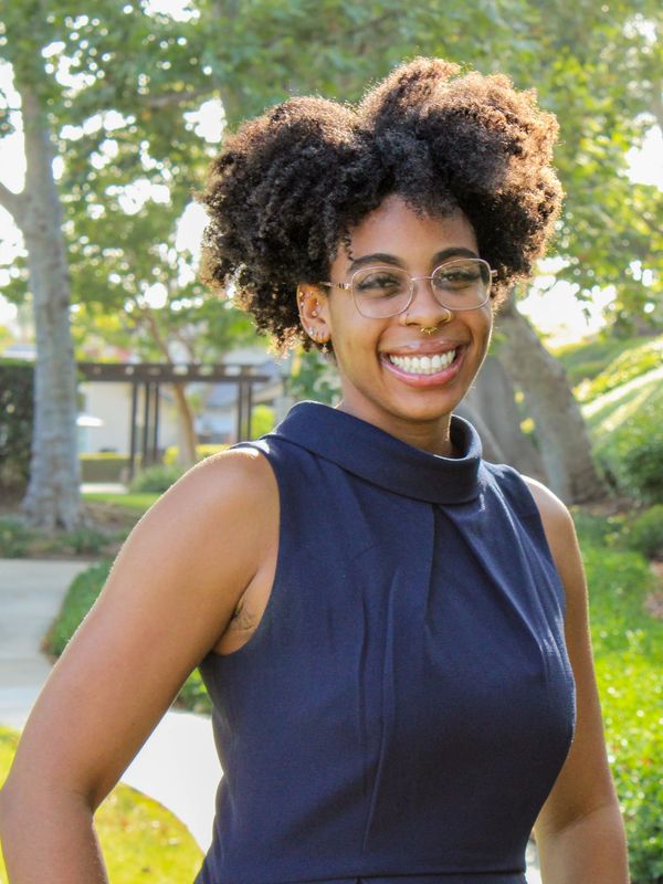 Smiling woman with curly hair and glasses stands outdoors on a sunny day.