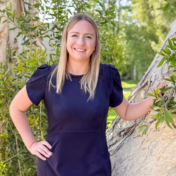 Smiling woman in a navy dress stands outdoors by a tree with greenery.