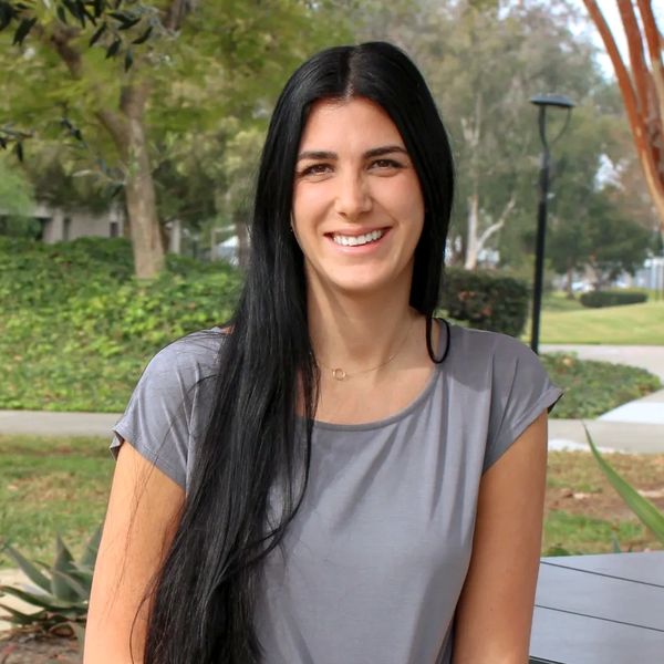Young woman with long black hair sitting on a bench in a park, smiling at the camera.