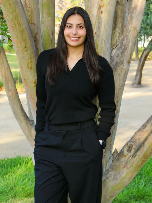 Young woman in black outfit standing and smiling by a tree outdoors.