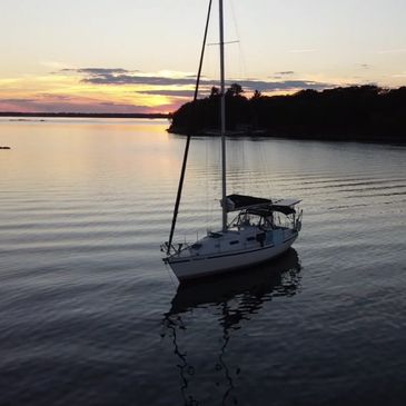 Sailboat anchored on calm water at sunset near a tree-lined shore.