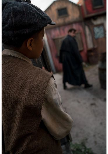 A young boy in vintage clothing watches a man walking past old buildings.