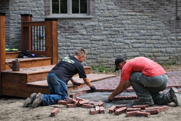 Two men laying bricks on the ground near a wooden deck.