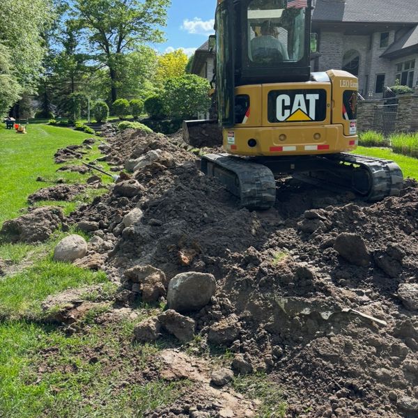 Excavator digging up soil near a large house on a sunny day.
