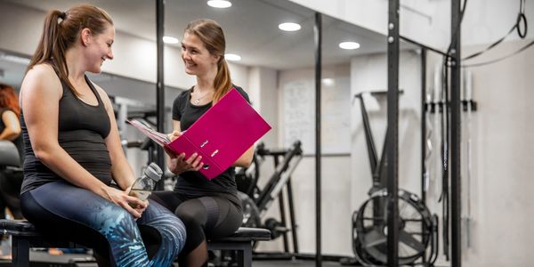 Two women chatting in a gym, one holding a water bottle, the other a pink folder.
