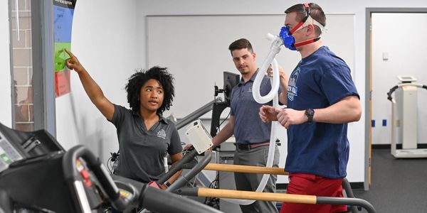 A man running on a treadmill wearing a respiratory mask with two trainers assisting.