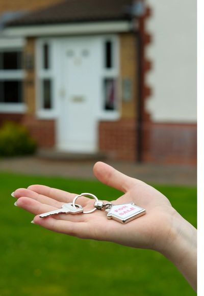 A hand held out with a key and keyring.  A blurred image of a new house in the background.