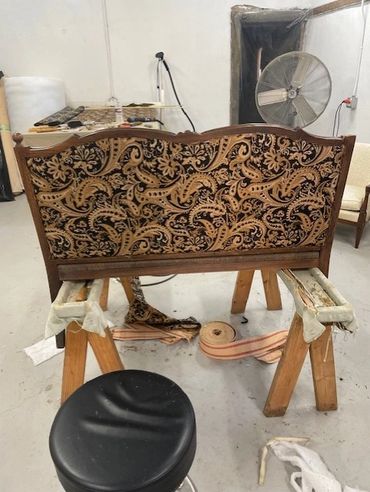 Antique wooden chair backrest with ornate fabric upholstery in a workshop.