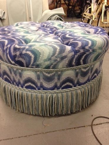 Round cushioned ottoman with blue patterned fabric and fringe trim.