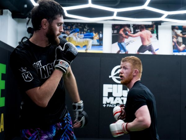 Two men spar in a modern MMA gym with gloves and focused expressions.